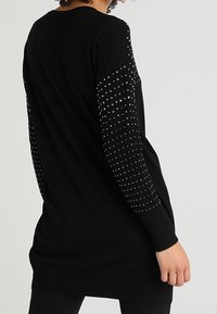 Femme portant une tunique noire à manches longues ornée de clous argentés sur les manches, debout devant un fond clair uni.