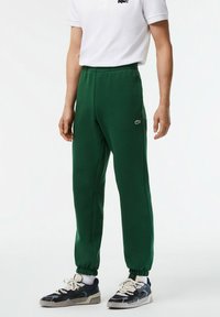 Pantalones de chándal verdes con cintura elástica y tobillos ajustados, con un logo blanco de Lacoste en el muslo izquierdo.