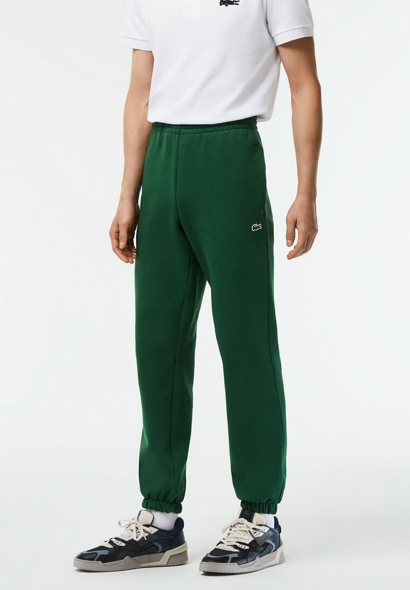 Pantalones de chándal verdes con cintura elástica y tobillos ajustados, con un logo blanco de Lacoste en el muslo izquierdo.