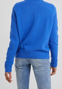 En person som bär en lysande blå stickad tröja och ljusblå jeans sedd bakifrån mot en enkel bakgrund.