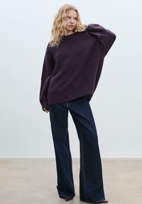 Überdimensionierter lila Strickpullover mit gerippten Bündchen, kombiniert mit dunkelblauen Schlagjeans mit auffälligen Nähten. Einheitlicher Hintergrund.