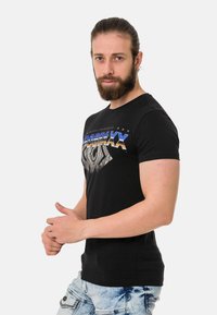 Schwarzes T-Shirt mit kurzen Ärmeln, featuring einem grafischen Design in Blau und Gold, Rundhalsausschnitt, aus weicher Baumwolle, kombiniert mit hellblauen Jeans.