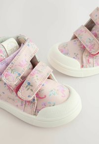 Lyserøde canvas sko med blomster mønstre, der har to velcro stropper, hvide gummisåler og teksturerede detaljer på tåen.