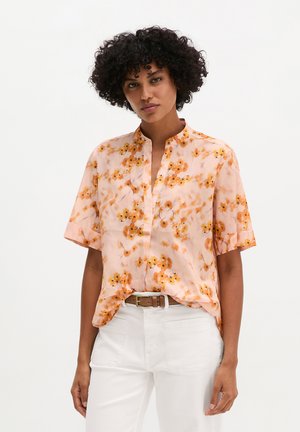 Kobieta o kręconych włosach, ubrana w brzoskwiniową, kwiecistą bluzkę z krótkim rękawem, wpuszczoną w białe spodnie z brązowym paskiem, stojąca na jednolitym tle.