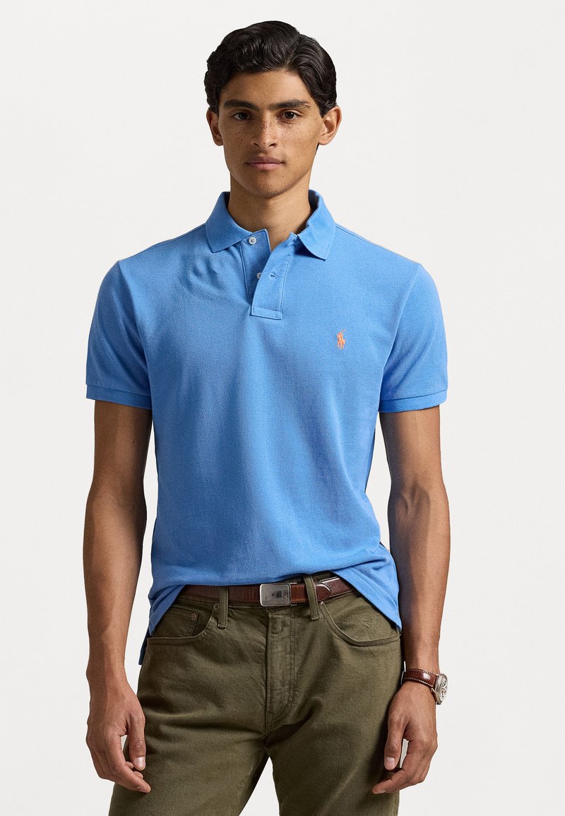 Camisa polo azul claro con un acabado texturizado, diseño con cuello y un logo naranja. Combinada con pantalones verde oliva y un cinturón marrón.