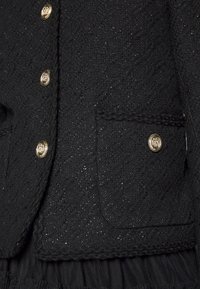 Černá texturovaná látková bunda s dekoračními zlatými knoflíky, přední kapsou a vlnkovým detailem podél spodního lemu.