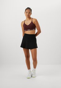 Sostén deportivo burdeos con banda blanca con el logo de Nike, combinado con una falda negra de diseño acampanado. La modelo lleva zapatillas deportivas blancas y verdes.