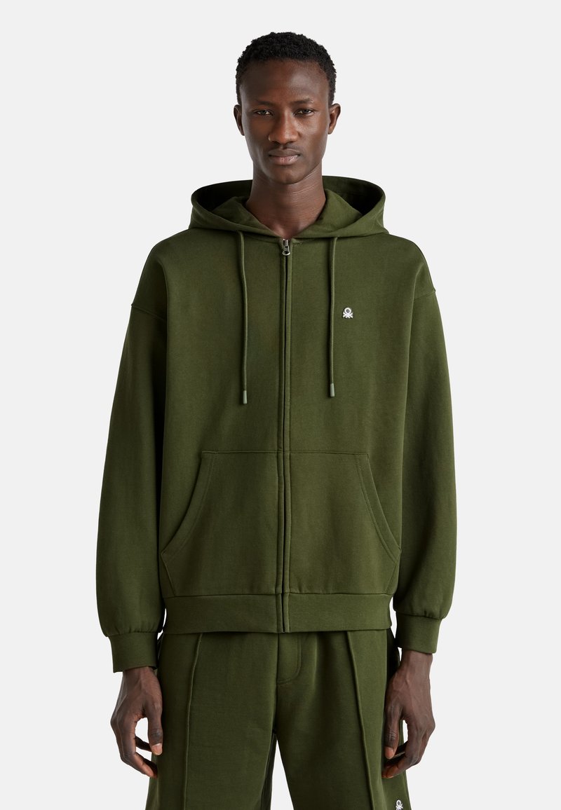 Felpa con zip color verde oliva realizzata in tessuto morbido, dotata di cappuccio regolabile con cordino, tasche frontali e un piccolo logo sul petto.