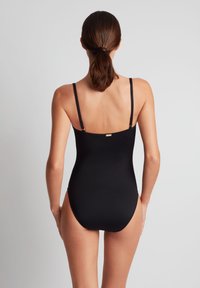 Kvinne med brunt hår i lav hestehale som har på seg en svart ett-delt badedrakt med tynne skulderstropper, sett bakfra.
