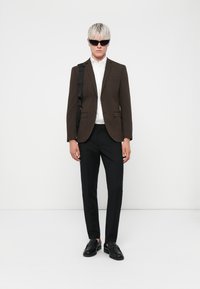 Sacou maro peste o cămașă albă, pantaloni negri și pantofi negri. Modelul poartă ochelari de soare negri și stă în fața unui fundal alb.