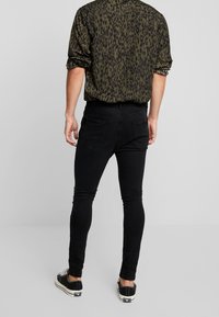 Jeans ajustados negros con una textura suave, combinados con una camisa de manga larga verde con estampado de leopardo y zapatillas negras. Bolsillos traseros visibles.