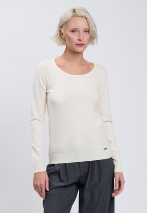 Femme aux cheveux blonds courts, portant un pull blanc uni à manches longues et un pantalon plissé gris foncé, debout devant un fond uni.