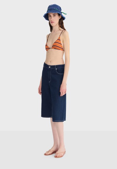 Femme portant un haut de bikini rayé orange, un short en denim foncé jusqu'aux genoux, un chapeau seau bleu et des tongs beiges, debout devant un fond neutre.