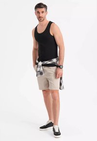 Svart tank top, beige shorts, rutig flanell skjortan knuten runt midjan, och svarta sneakers med vita sulor. Enkelt bakgrund, modell stående upprätt.