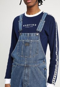 Pantalones de peto de mezclilla azul con tirantes ajustables, con un bolsillo delantero y una etiqueta de logo, combinados con una camisa de manga larga azul marino con detalles en blanco.
