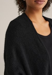 Sciarpa nera lavorata a maglia con trama testurizzata, che cade sopra una maglia nera. Il tessuto appare morbido e accogliente, caratterizzato da un design con apertura frontale.