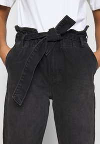 Pantalons en denim noir à taille haute, avec une ceinture à nouer et des poches latérales. Le tissu a une finition douce et texturée.