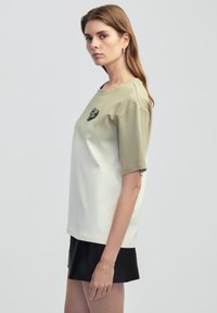 T-shirt à manches courtes avec un design dégradé dans des tons verdâtres et blanc cassé, rehaussé d'un cœur noir sur la poitrine.
