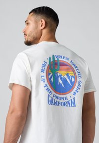 Witte katoenen t-shirt met een kleurraf rond ontwerp van een cactus, bergen en tekst "WANNEER DE NATUUR BELLEN, NEEM DE TELEFOON OP CALIFORNIË."