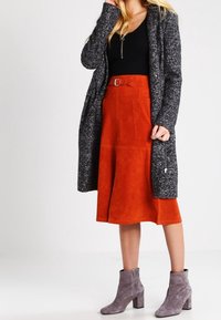 Nainen, joka on pukeutunut mustaan toppiin, pitkään tummanharmaaseen neuletakkimekkoon, ruosteenväriseen mokkahiihdon midi-hameeseen vyöllä ja harmaisiin mokkanahkasaappaisiin, yksinkertaista taustaa vasten.