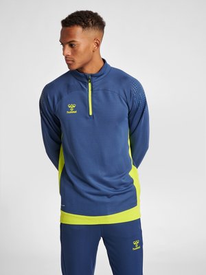 Mężczyzna w niebiesko-neonowożółtym stroju sportowym z półzamkiem i pasującymi spodniami, stojący z rękami za plecami, patrzący w lewo.