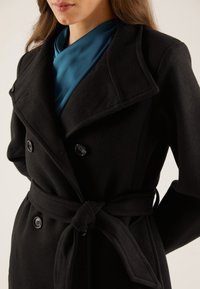 Černý kabát z vlněné směsi s dvouřadým zapínáním, velkými klopami a vázačkou v pase. Pod ním je vidět tyrkysová saténová blůza.