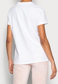 Person i en enfärgad vit kortärmad t-shirt och ljusrosa leggings med subtila vita Nike-logotyper, stående mot en enkel bakgrund.