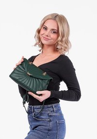 Zelená kožená kabelka s prošívaným designem, zlatými akcenty a klopovým uzávěrem; drží ji osoba v černém topu a džínech.