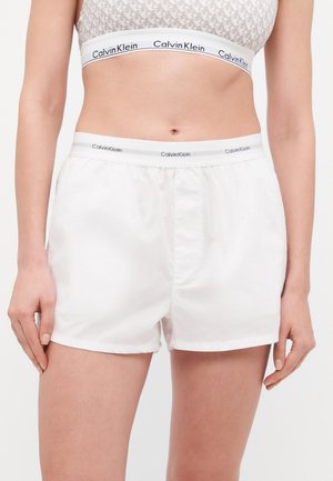 White cotton shorts with an elastic Calvin Klein waistband, featuring a smooth texture and a relaxed fit. Minimalistic design, knee-length.