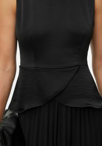 Gros plan sur un haut noir sans manches avec un détail taille peplum porté sur une jupe plissée, porté par une personne tenant un sac noir texturé.