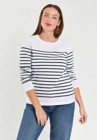 Hvid langærmet skjorte med marineblå vandrette striber, rund halsudskæring og subtilt brandlogo på kanten. Parret med blå jeans.