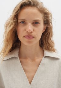 Camicia di lino beige chiaro con colletto e scollo a V, caratterizzata da un tessuto texturizzato e una vestibilità comoda, indossata da una persona con capelli mossi.