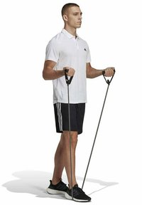 Witte polo-shirt, zwarte shorts met drie strepen, houdt weerstandsbanden vast met zwarte handgrepen. Donkere sport schoenen met een lichtgewicht ontwerp.