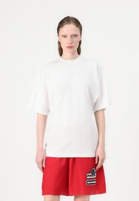 Witte oversized T-shirt met korte, verlaagde mouwen en een ronde halslijn, gedragen met rode shorts met zwart-witte geborduurde patches.