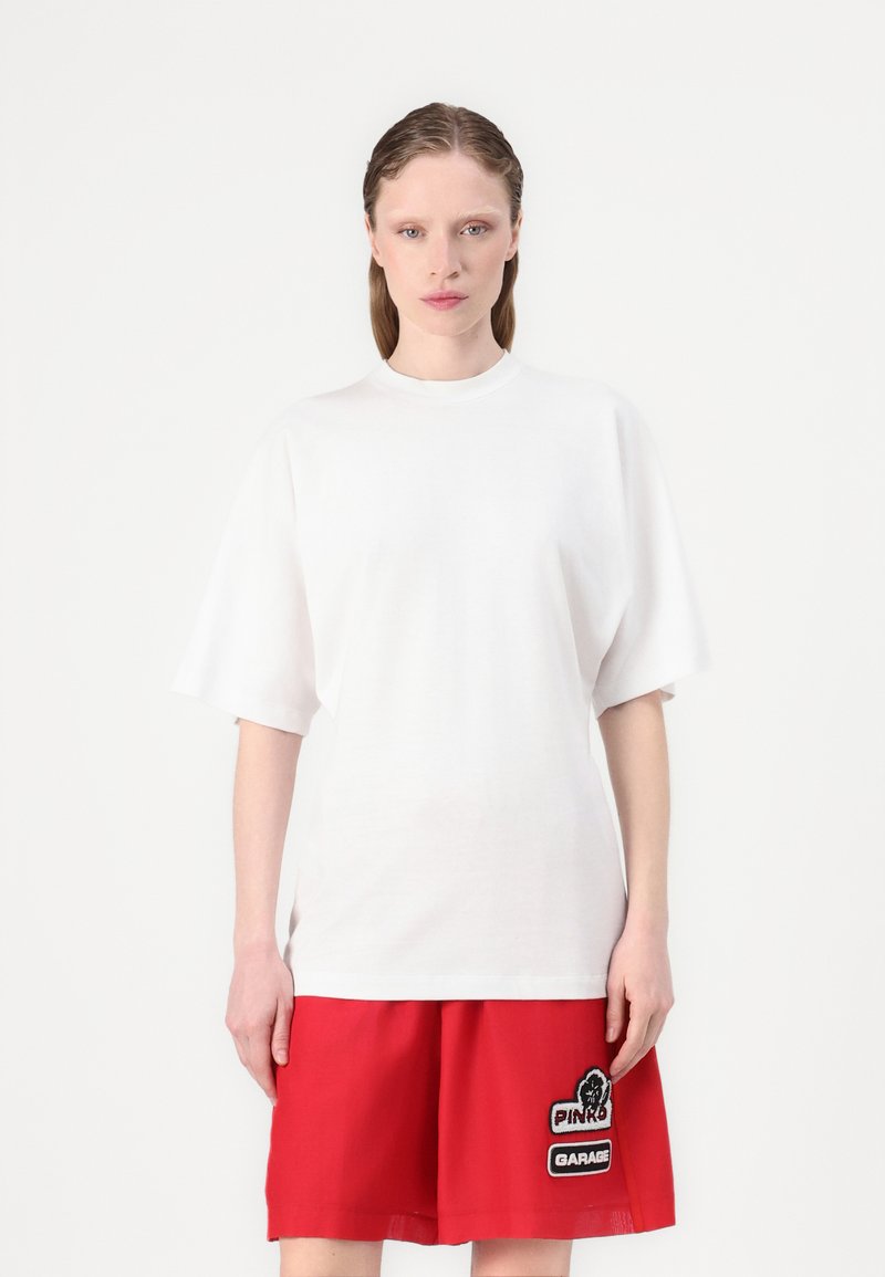 Witte oversized T-shirt met korte, verlaagde mouwen en een ronde halslijn, gedragen met rode shorts met zwart-witte geborduurde patches.