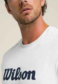 Hombre con barba que lleva una camiseta blanca con el logo "Wilson" en letras mayúsculas de color azul marino y un collar delgado de plata.