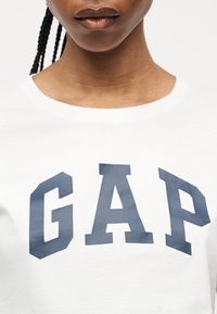 Femme portant un t-shirt blanc à col rond avec de grandes lettres "GAP" bleu marine sur la poitrine, cheveux tressés visibles sur le côté.