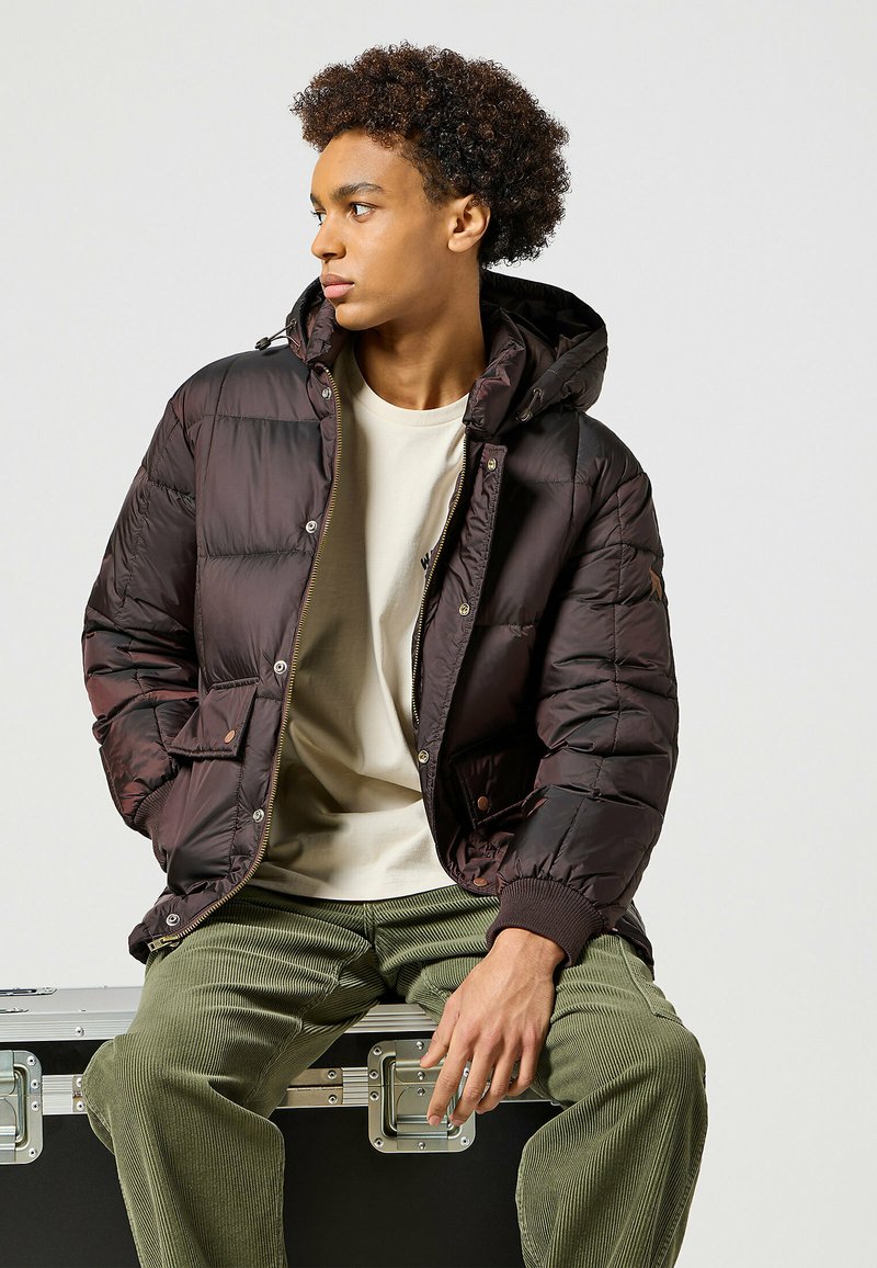 Giacca a vento marrone con cappuccio, finitura lucida, zip frontale e bottoni a pressione; indossata sopra una maglietta crema e pantaloni in velluto a coste verde oliva.