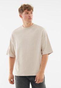 Tricou oversized bej din material moale, cu decolteu rotund și mâneci scurte, prezentat pe un model cu păr creț.