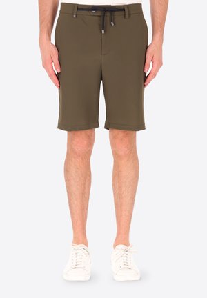 Uomo con pantaloncini casual verde oliva con coulisse nera in vita e sneakers bianche, in piedi davanti a uno sfondo semplice.