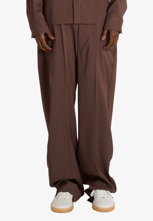 Persona che indossa pantaloni marroni larghi e sneakers bianchi con suole in gomma, in piedi davanti a uno sfondo chiaro e uniforme.