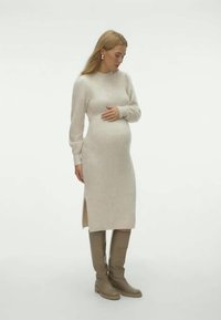 Kelio ilgio, kreminė megzta suknelė su ilgomis rankovėmis, šoninėmis plyšiais ir laisvu siluetu. Derinama su taupe spalvos kelių ilgio botais. Lygus paviršius.