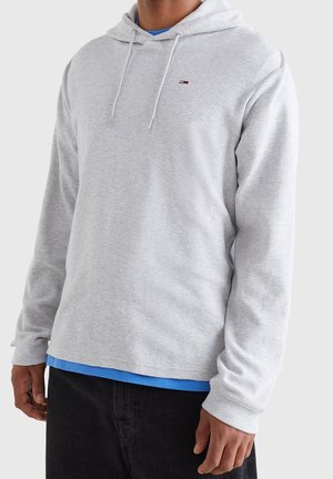 Hombre con sudadera de punto tipo waffle gris claro sobre camisa azul y pantalones negros, de pie contra un fondo gris liso.