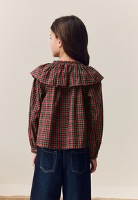 Chemise à carreaux rouges et verts avec col à volants et manches longues, associée à un pantalon en denim bleu foncé à jambes larges. Vue de dos.