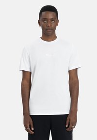 Witte katoenen t-shirt met een ronde hals, korte mouwen en een subtiel logo in het midden. Gladde textuur en klassieke pasvorm, gecombineerd met zwarte broek.