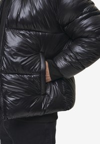 Sort pufferjakke med en blank, quiltet tekstur, der har en lynlåslukning og ribbede manchetter, vist med en hånd i en sidelomme.
