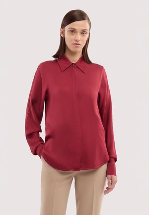 Vrouw met recht bruin haar, gekleed in een rood blouse met lange mouwen en beige broek, staande tegen een effen lichte achtergrond.