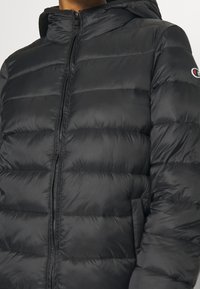 Svart vadderad puffjacka med huva, framdragkedja och liten logopatch på överarmen, bärs av en person.