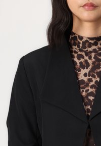 Černý blazer s širokým límcem, navržený s hladkým povrchem. Pod ním je vidět hnědý top s vysokým límcem a leopardím vzorem.