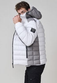 Giacca imbottita bianca con accenti grigi, dotata di cappuccio, tasche con zip e tessuto testurizzato. Vestibilità ampia con design a righe orizzontali.